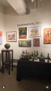 Art depicting and inspired by the natural world hangs at a gallery showing, the words "Environmental Ties II" on the wall above several paintings. A table of beverages is in the foreground.
